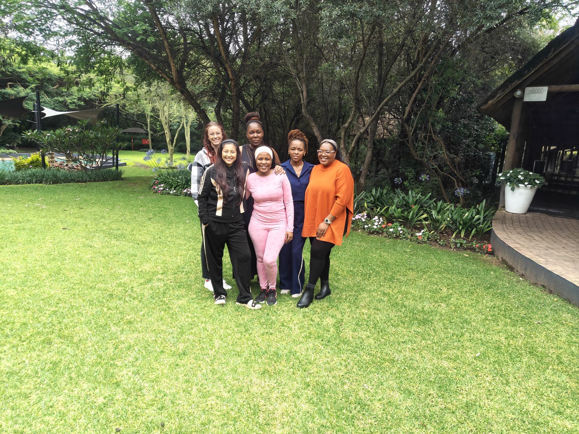 Tricia, Nikki, Ziznisa, Phozisa, Sandyand Lucia stand smiling together on a lush green lawn in front of trees and a patio.