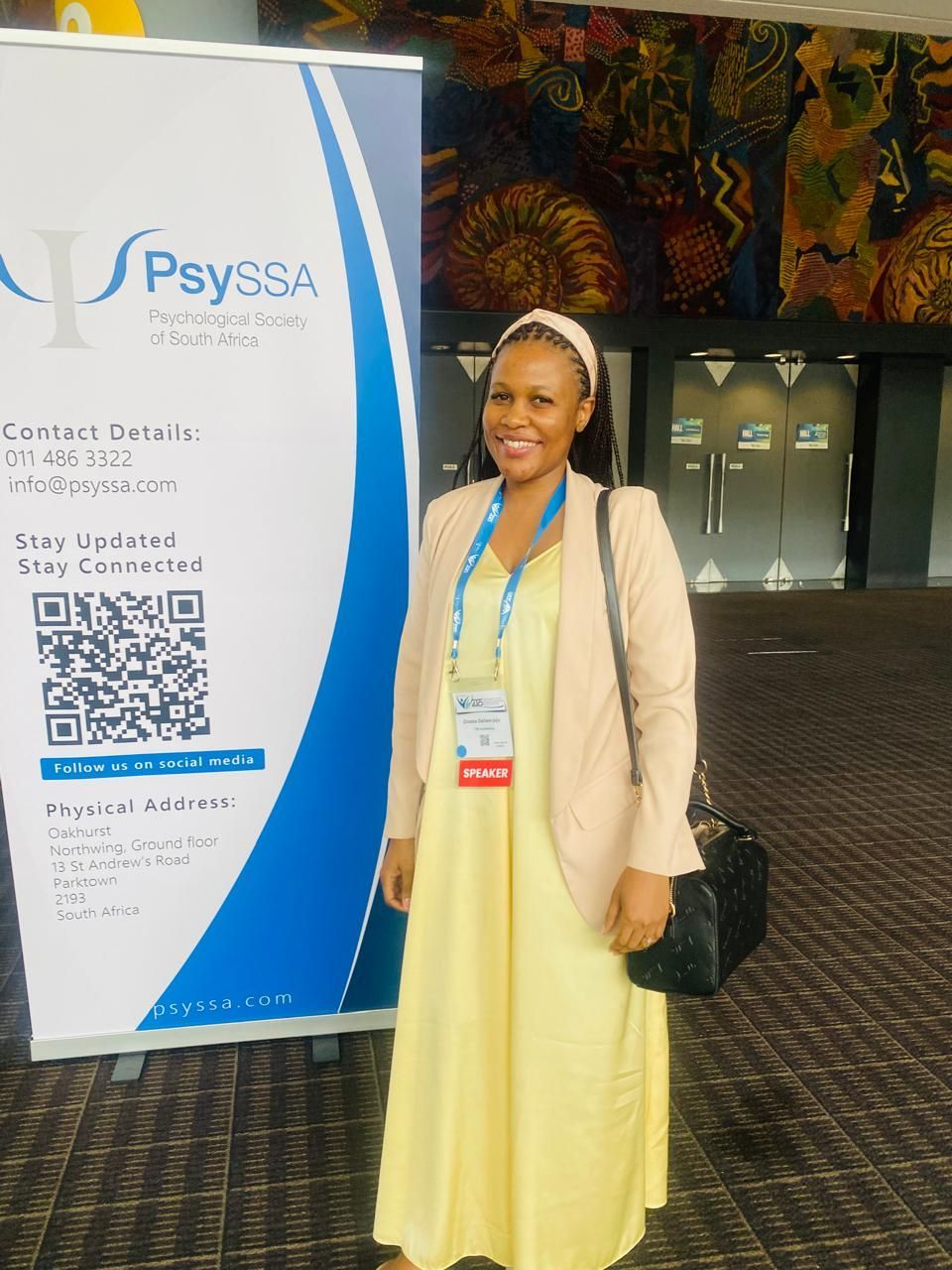 Zinzisa smiles while standing next to a vertical PsySSA banner at an indoor event, wearing a yellow dress and beige blazer.