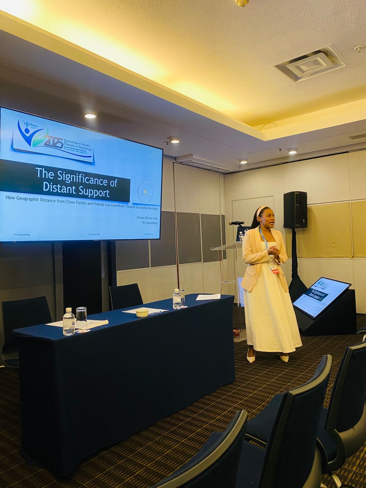 Zinzisa in a light blazer and skirt stands near a projector screen in a meeting room, presenting to an audience.
