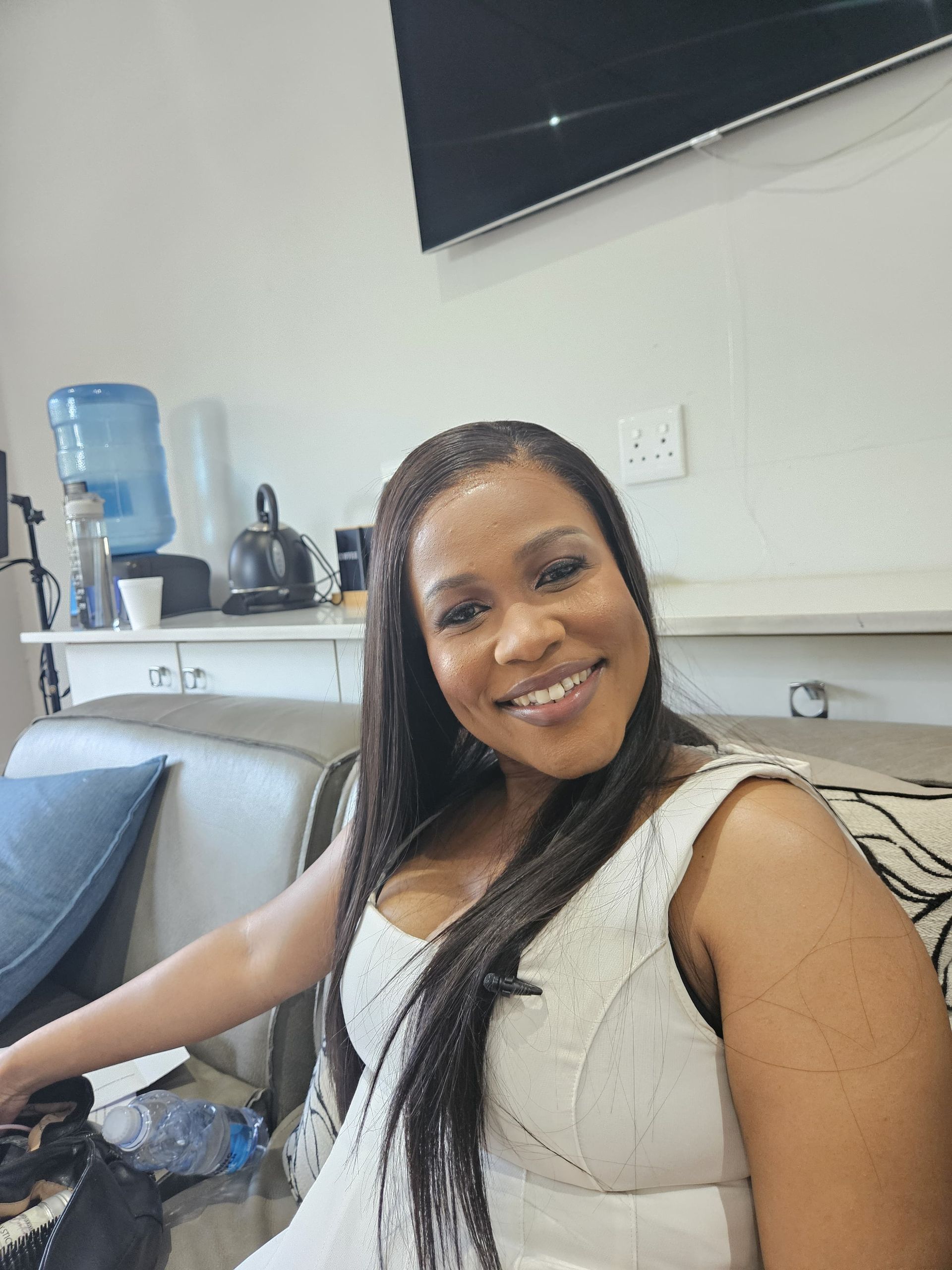 Zinzisa with braided hair and a white sleeveless dress smiles while sitting on a couch in a room with a water dispenser.