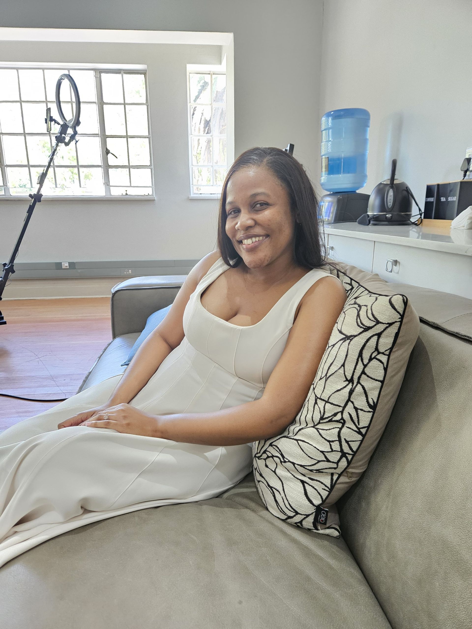 Zinzisa Deliwe-Juju in a white dress sitting on a grey couch, smiling, in a room with a ring light and water dispenser.