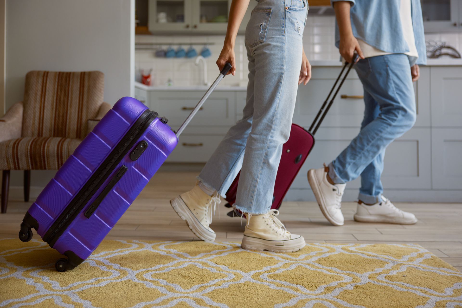 Two people rolling suitcases across a patterned rug in a bright room, one purple and one maroon.