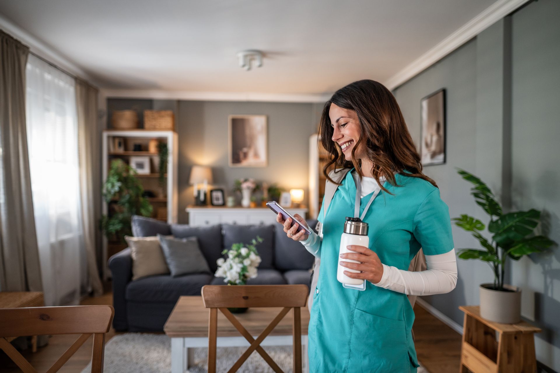 Woman in teal scrubs checking her phone in a cozy living room, holding a water bottle.