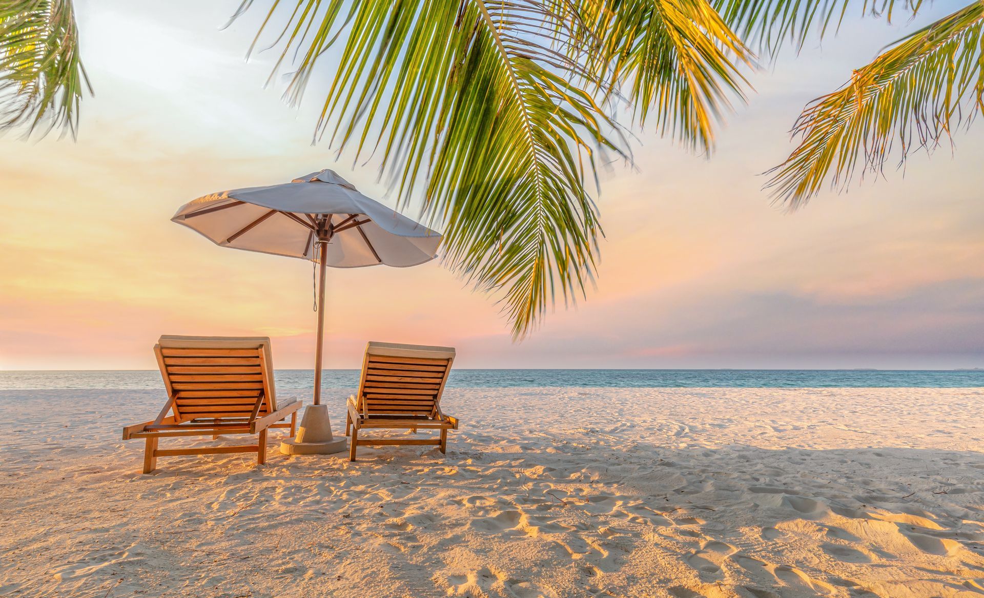 Two lounge chairs under a beach umbrella on a sandy beach at sunset, with palm fronds overhead.
