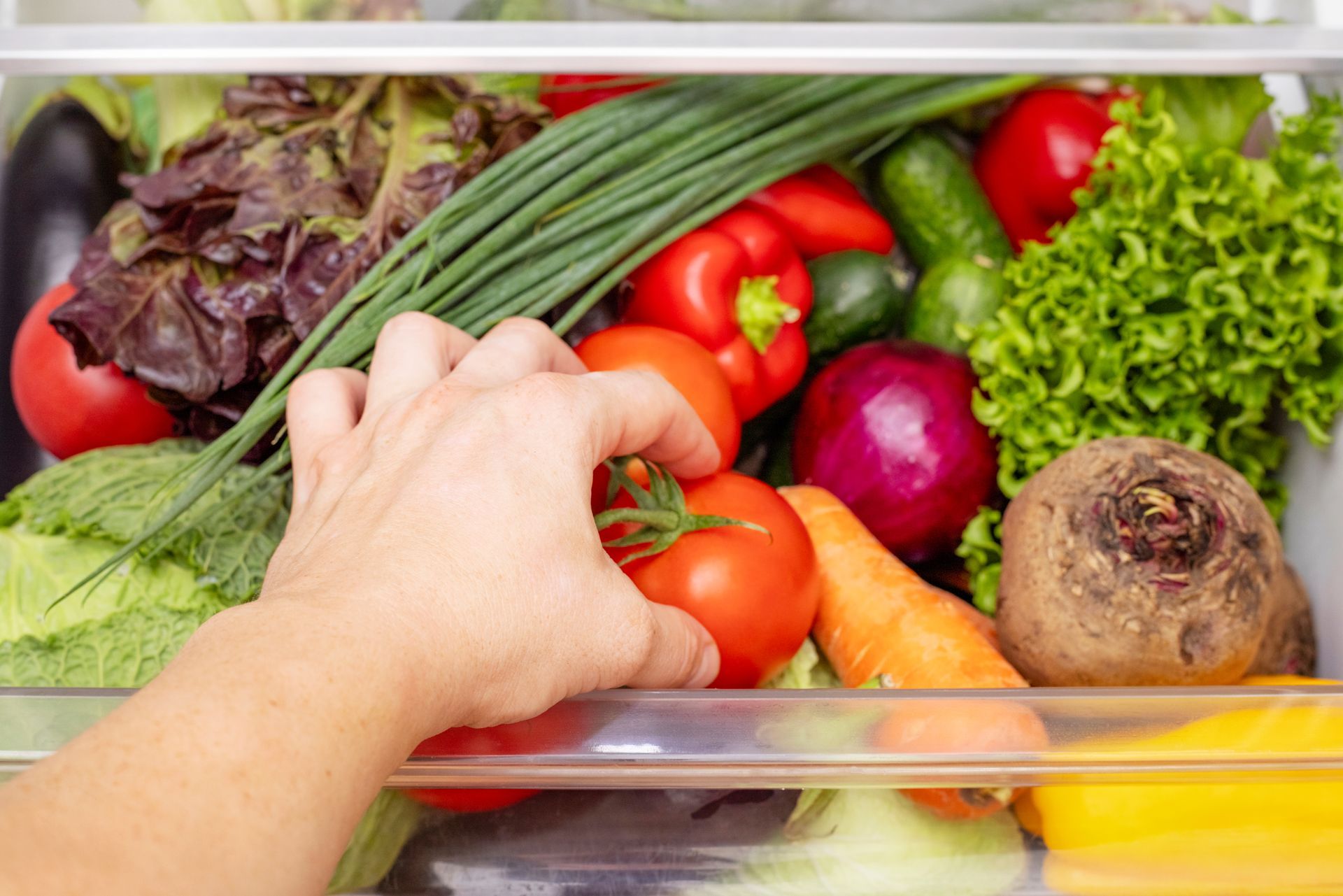 Hand reaching into a refrigerator drawer of fresh vegetables, including tomatoes, greens, onions, and a beet