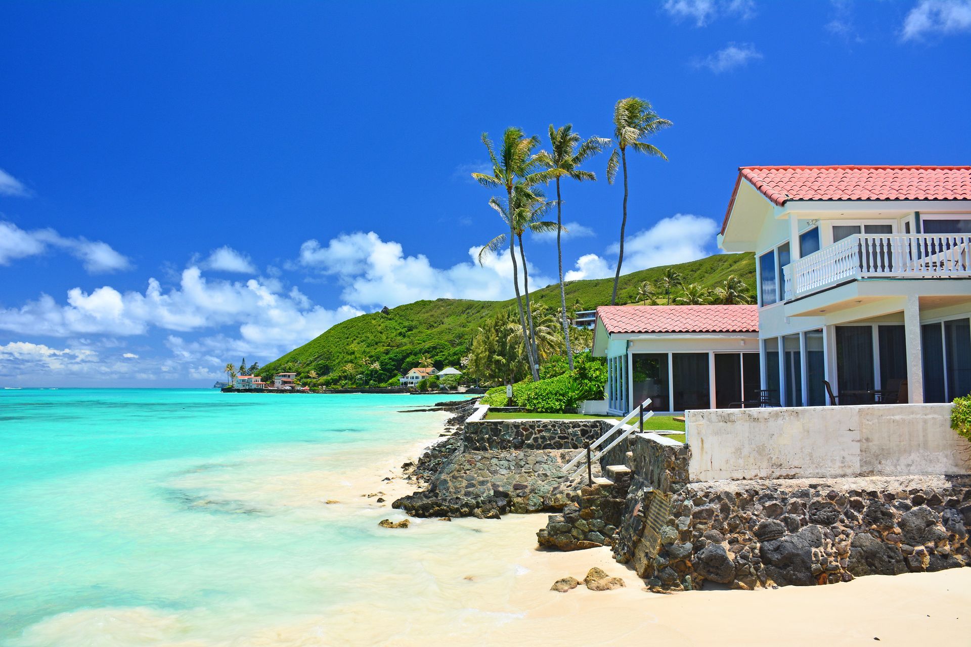 Tropical beach with turquoise water, palm trees, and a white house on a rocky shore under a blue sky