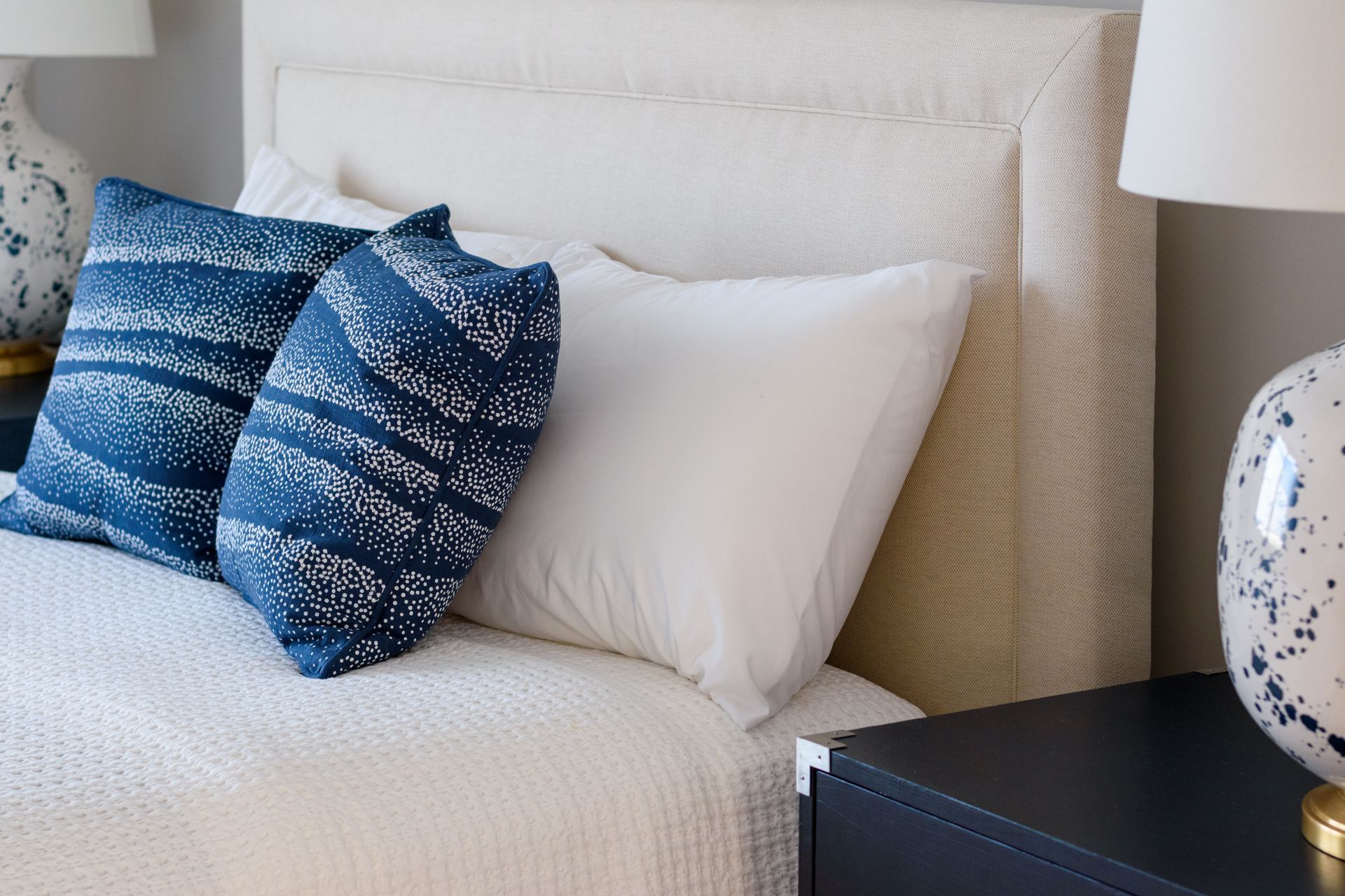 White bed with white pillows and blue patterned cushions beside a black nightstand and lamp