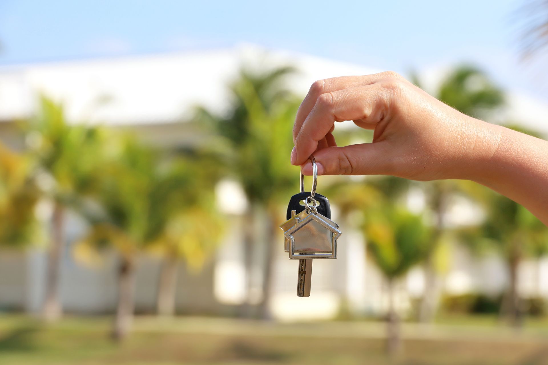 Hand holding house keys in front of a blurred suburban home and palm trees