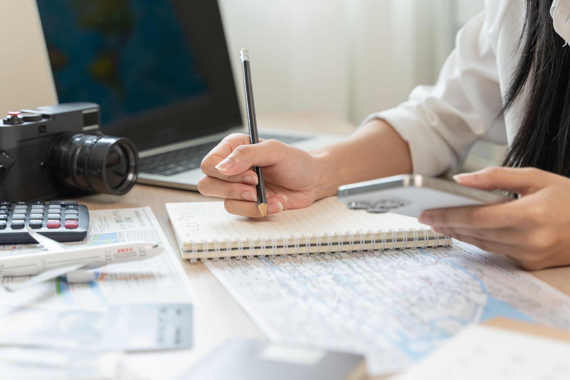 Person writing in a notebook beside a camera, calculator, and laptop on a desk