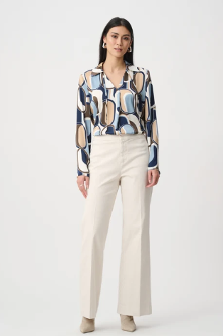Una persona con un blazer color cammello e pantaloni abbinati, con una maglia bianca testurizzata, è appoggiata a uno stipite di una porta nella luce tenue del sole.
