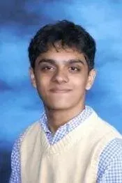 Portrait of a young person smiling, wearing a cream sweater vest over a checkered shirt against a blue backdrop