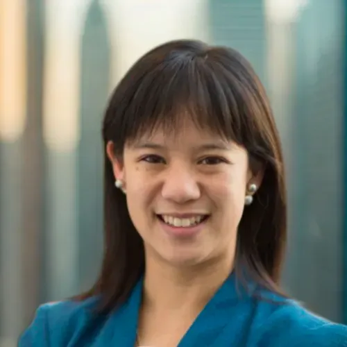 Smiling person in a blue blazer, standing against a blurred outdoor city background