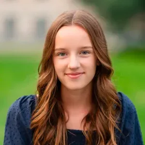 Portrait of a smiling person with long brown hair, standing outdoors against a blurred green background