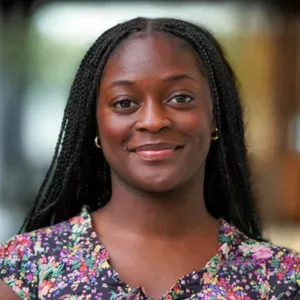 Smiling woman with braided hair in a floral blouse, standing outdoors with a blurred background