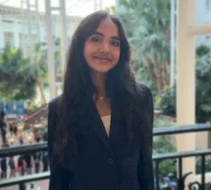 Smiling woman in a dark blazer standing on a balcony with a blurred indoor atrium behind her
