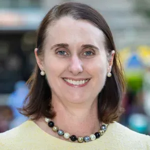 Smiling woman in a yellow top and beaded necklace against a blurred outdoor background