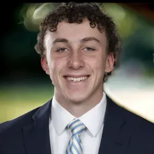 Smiling person in a navy suit and striped tie against a blurred green background