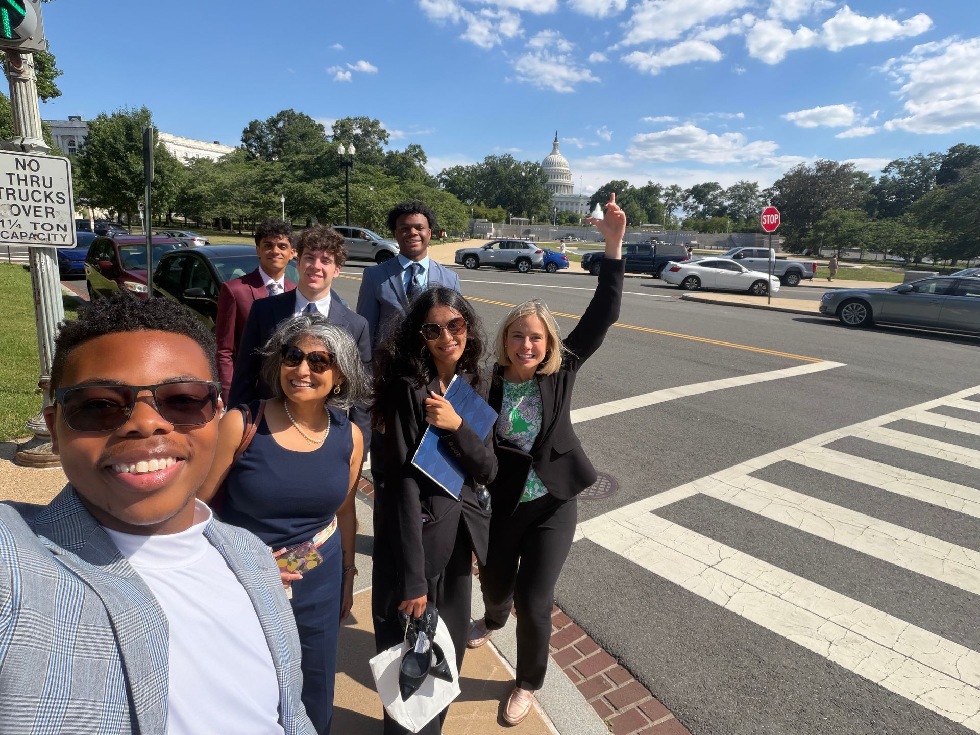 Group of five smiling friends posing at a crosswalk on a sunny city street, one holding a phone aloft.