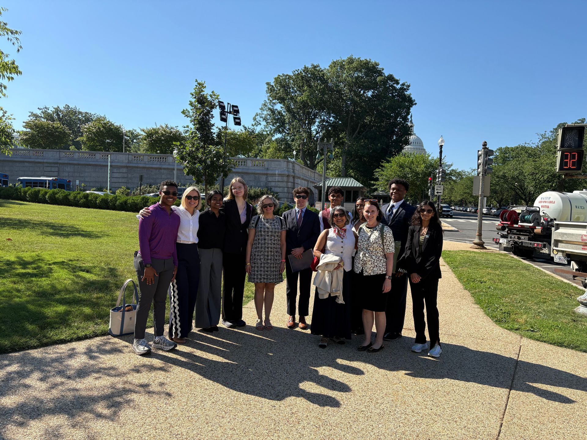 Group of people posing on a sunny sidewalk beside a grassy hill and road, with cars and trees in the background