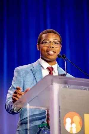 Speaker at a podium with microphone, wearing a light blue suit against a blue curtain backdrop.