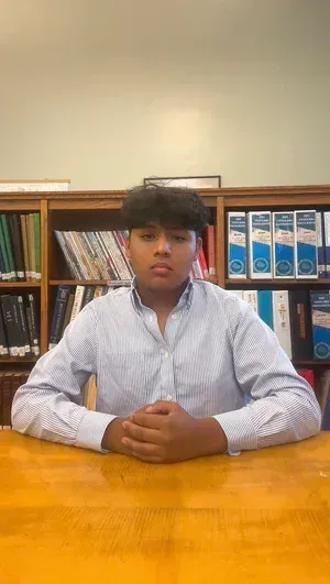 Person seated at a wooden table in a library, hands clasped, with bookshelves in the background.