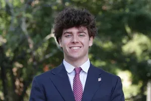 Smiling person in a blue blazer, standing against a blurred outdoor city background