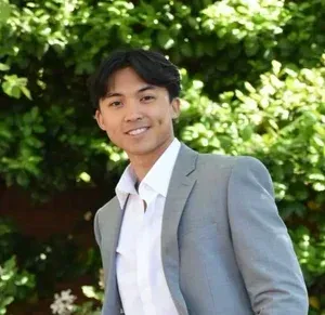 Smiling person in a light gray suit and white shirt standing outdoors against green foliage