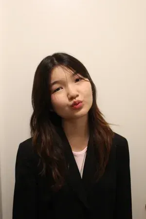 Person in a black blazer making a slight pout against a plain beige wall.