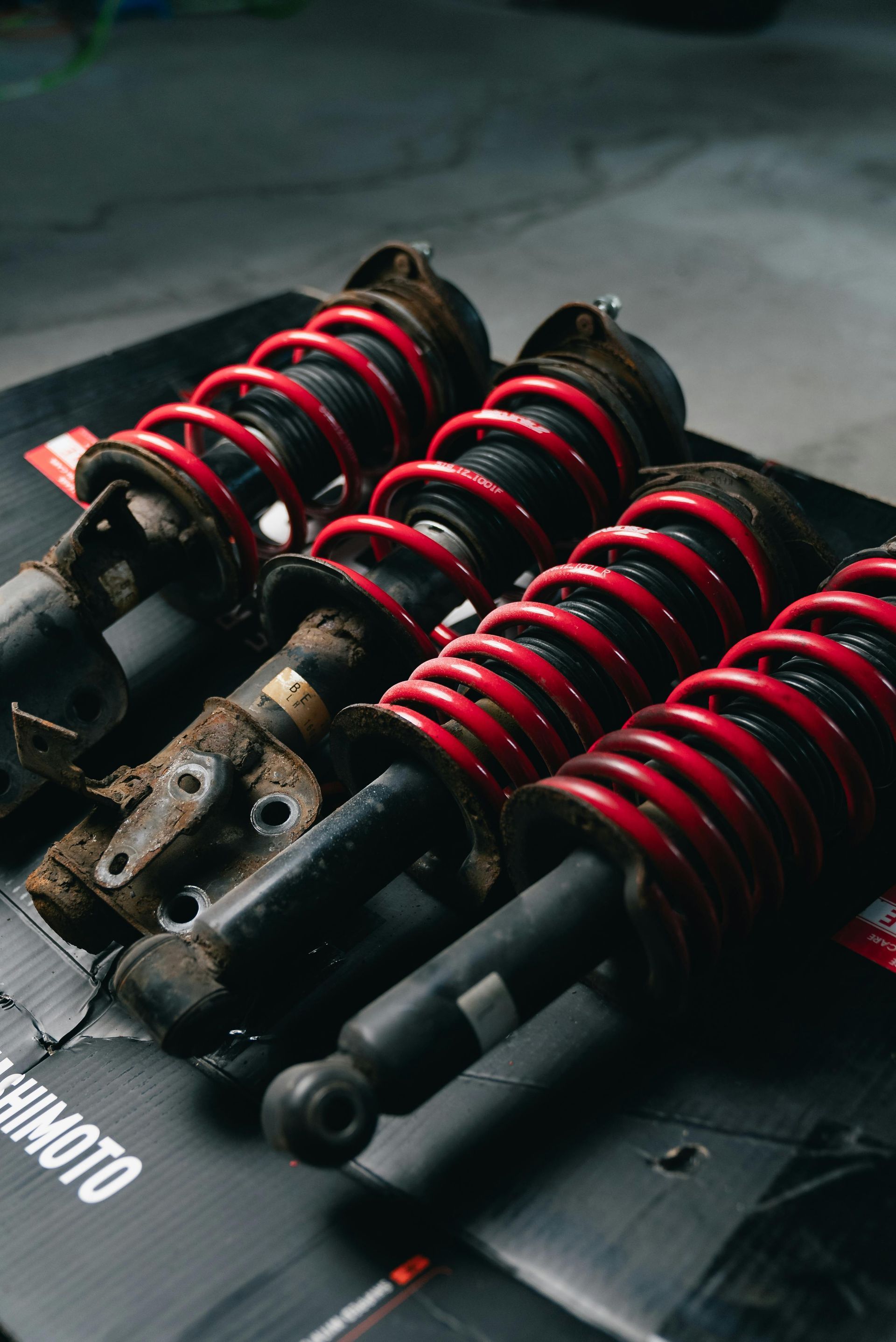 Four automotive shock absorbers with red springs, resting on a black surface.