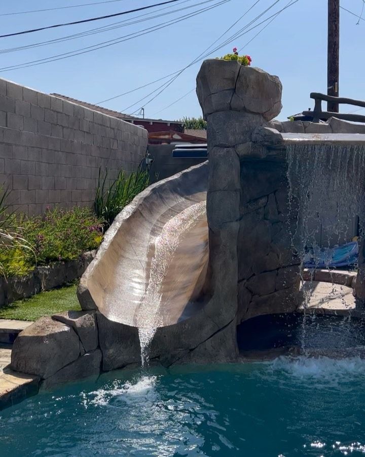 Water slide descending from a rock structure into a blue pool, with a person on top and cloudy sky above
