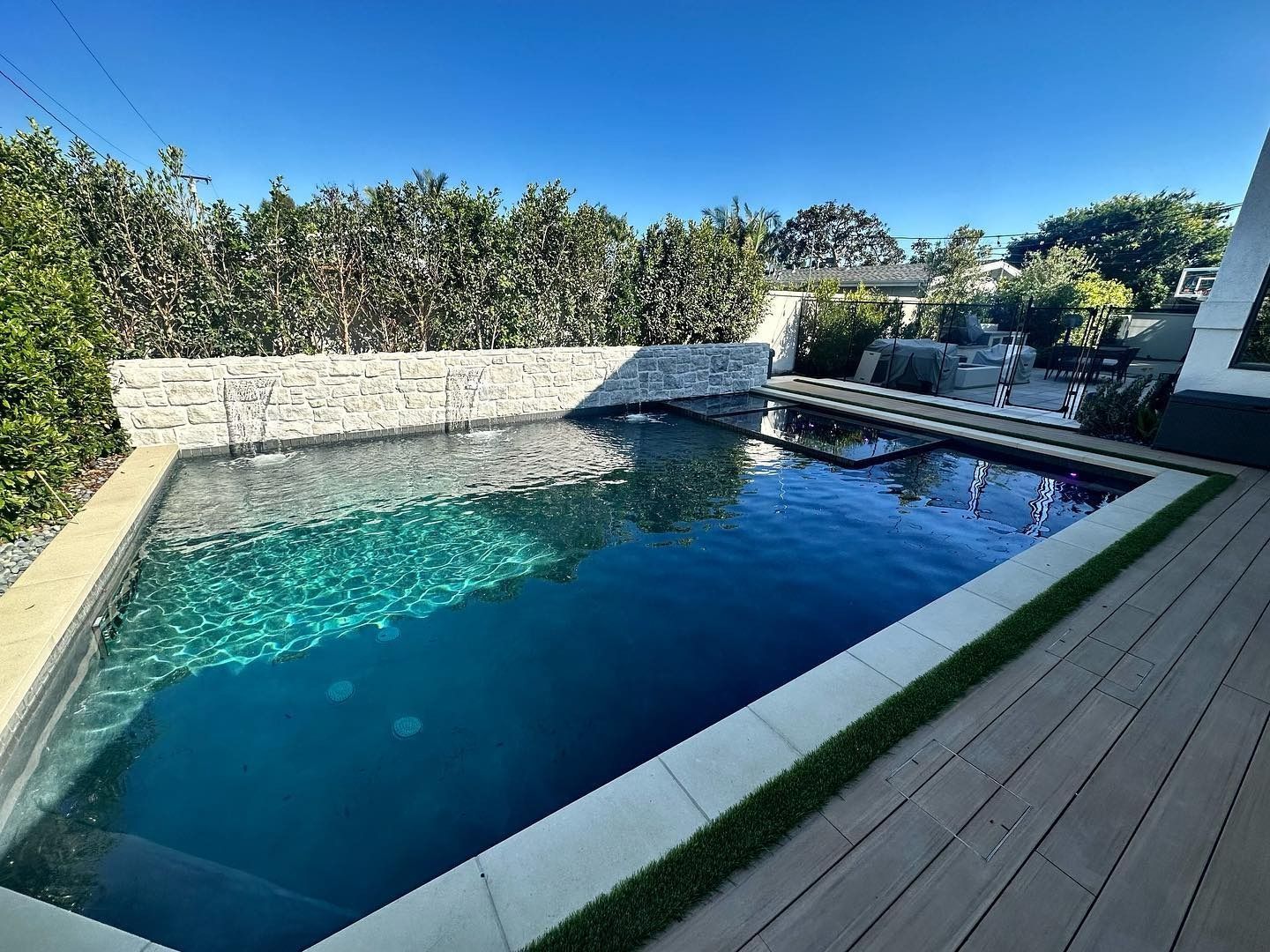 Modern backyard swimming pool with stone wall, blue water, and wooden deck under a sunny sky