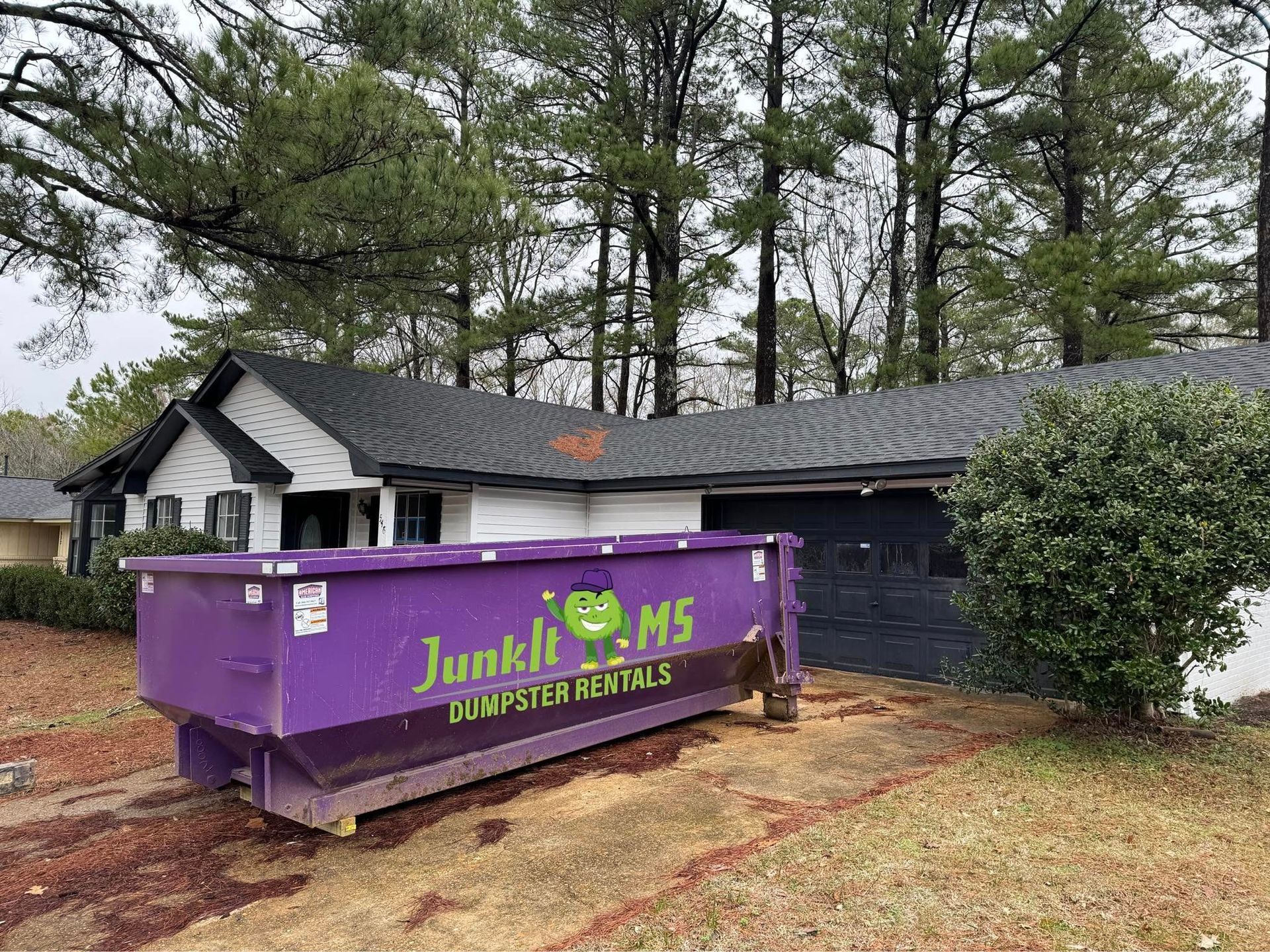 Purple dumpster in front of a house with a black roof and green shrubs, possibly for renovation.