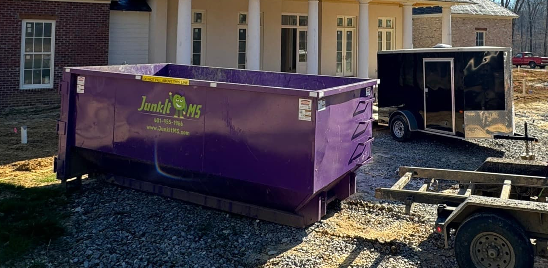 A purple dumpster in front of a house under construction, next to a black trailer.