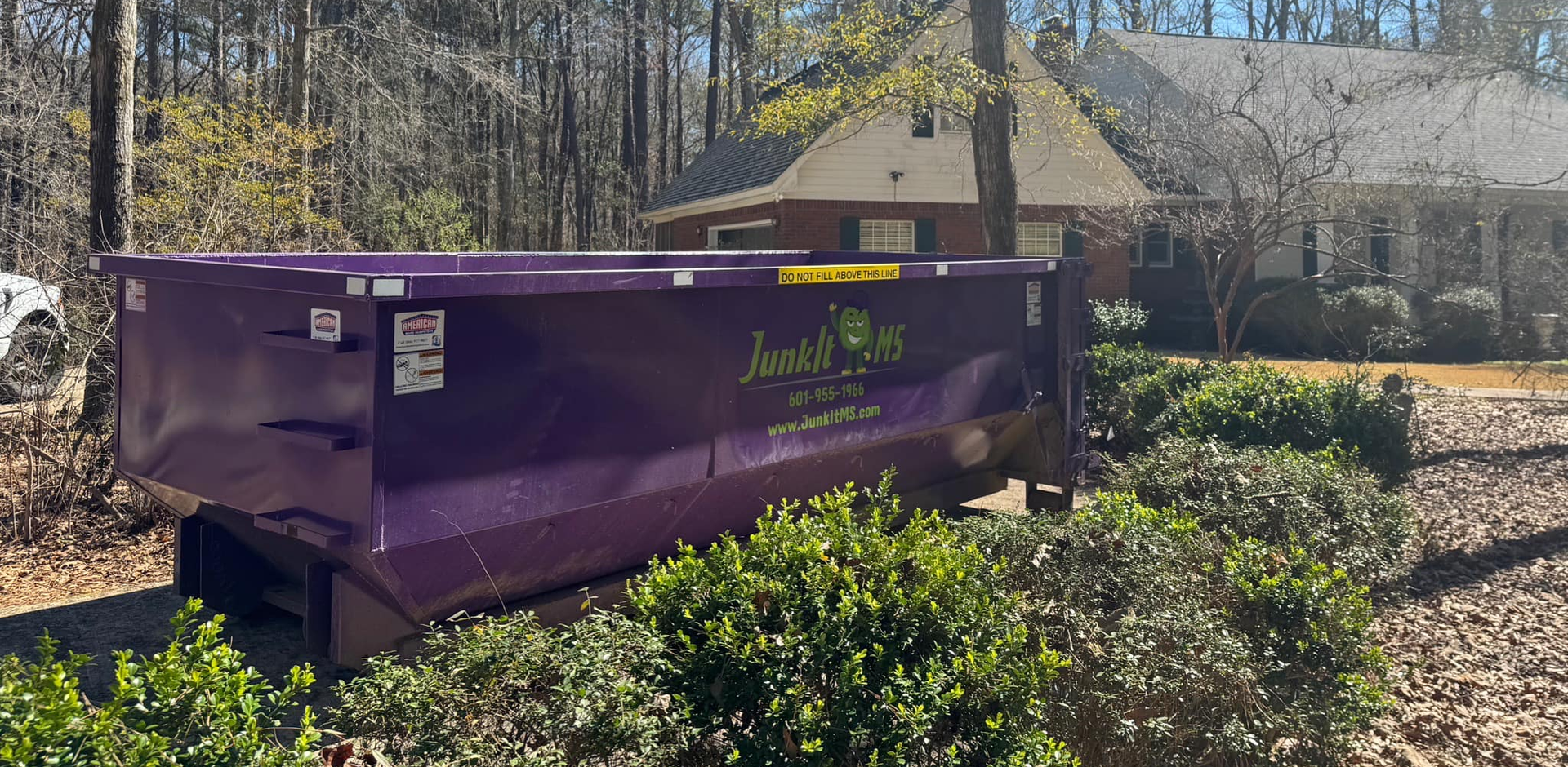 A purple dumpster in front of a house, surrounded by bushes, trees, and blue sky.