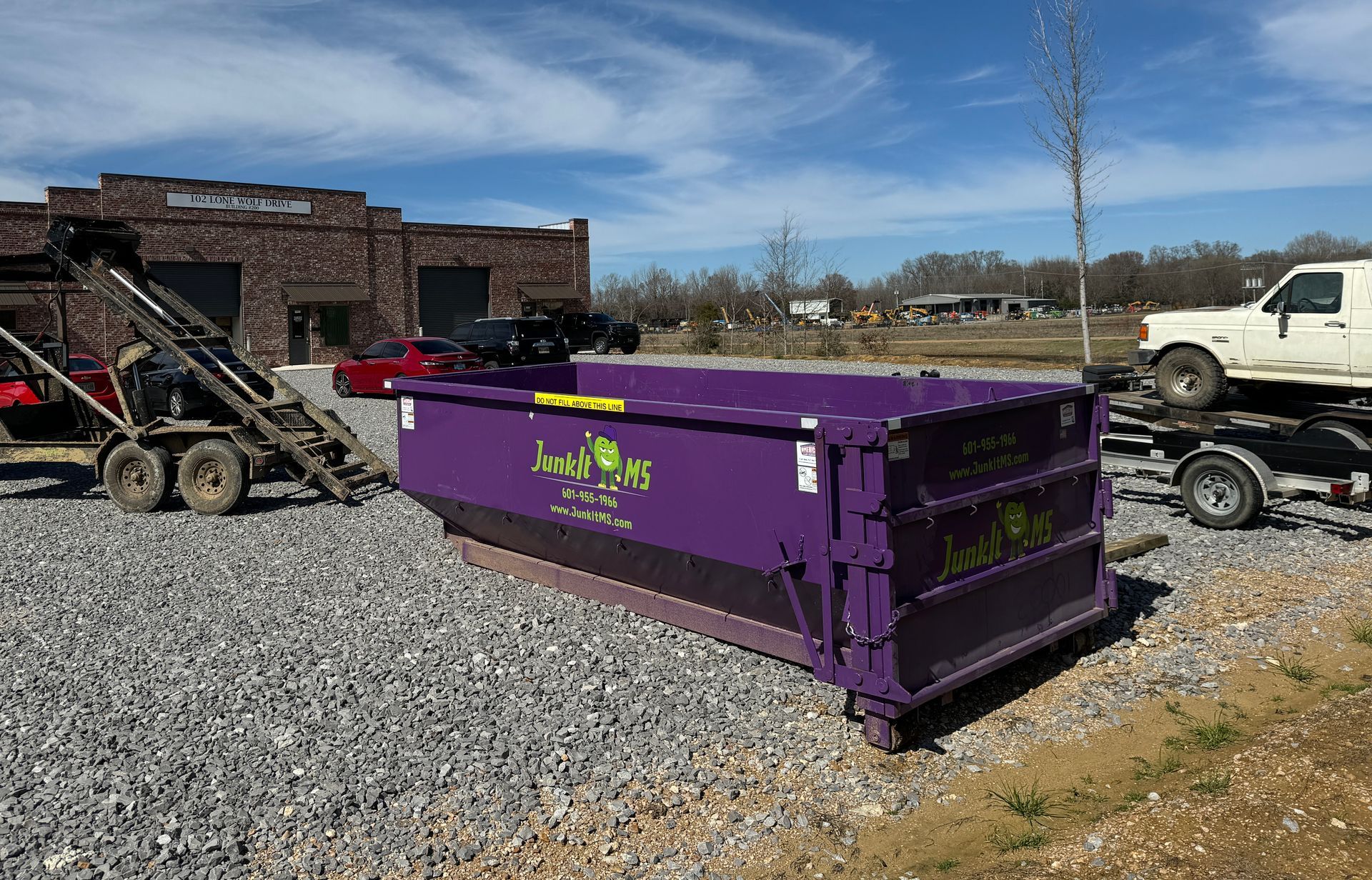 Purple dumpster on gravel, in front of a brick building and a truck on a trailer.