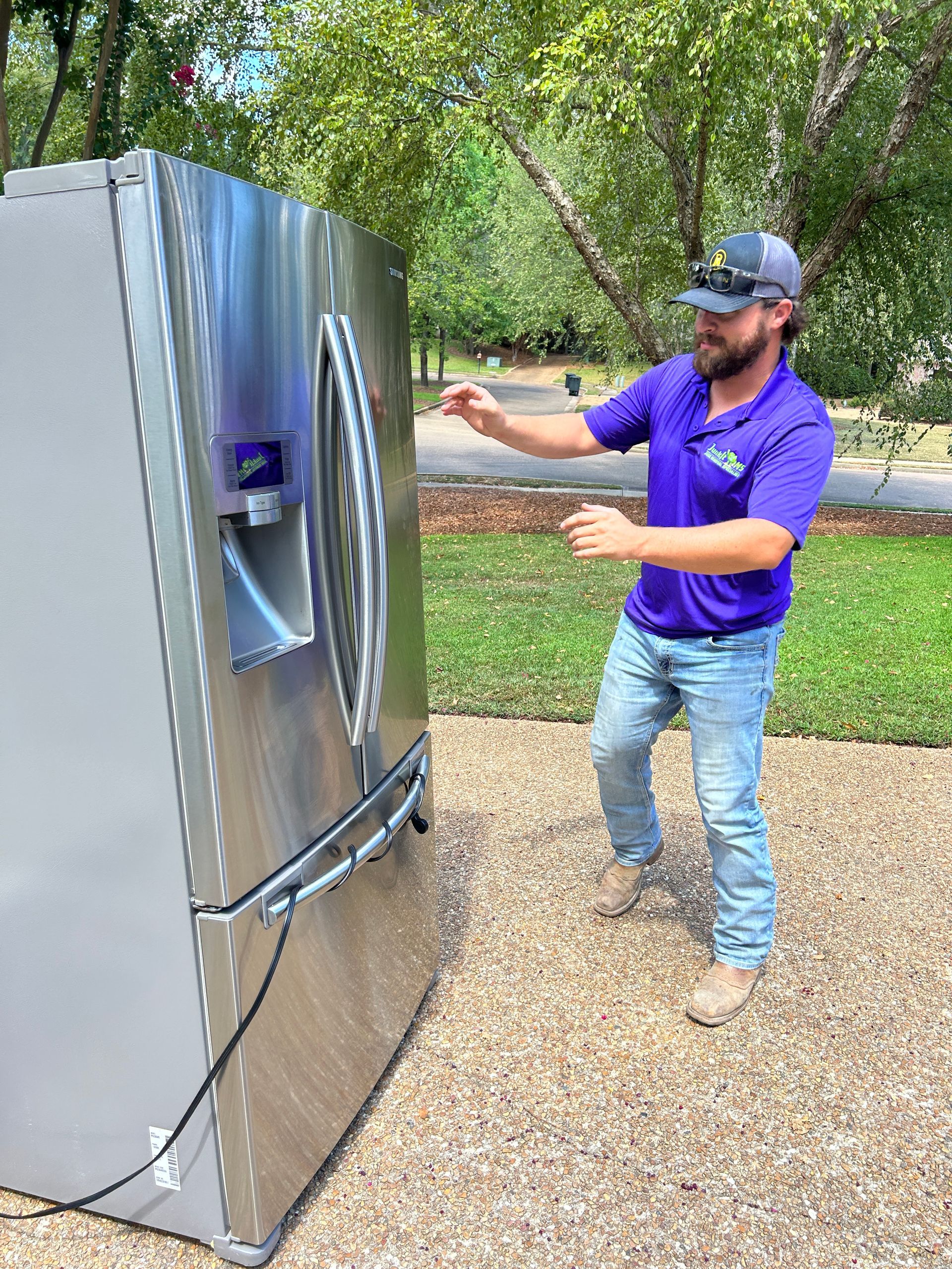 Man in purple shirt and jeans pushing a stainless steel refrigerator outside on a sunny day.