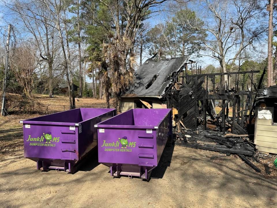 Two purple dumpsters in front of a charred, burned-down house on a sunny day.