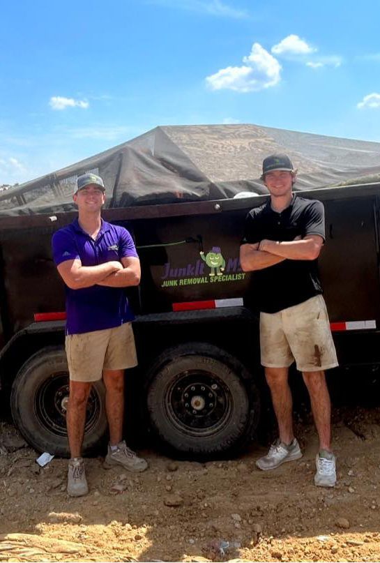 Two young men in work clothes stand with arms crossed in front of a black trailer on a sunny day.