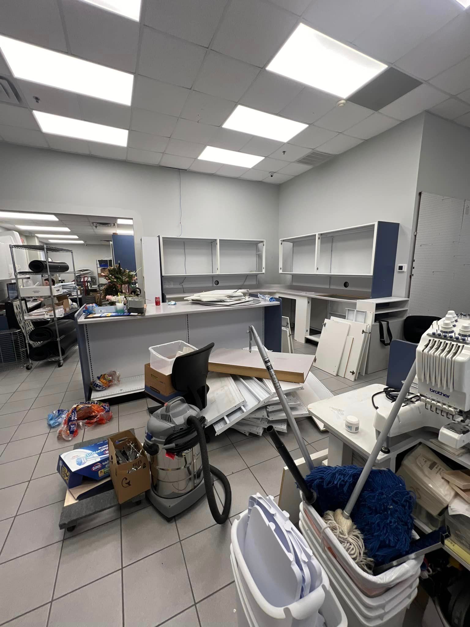 Messy room with cabinets, cleaning supplies, and construction materials, likely an office or lab undergoing renovation.