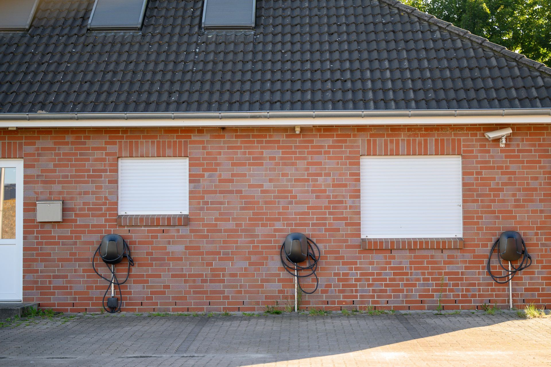 Un bâtiment en brique rouge avec trois bornes de recharge pour véhicules électriques fixées au mur et un toit noir avec panne