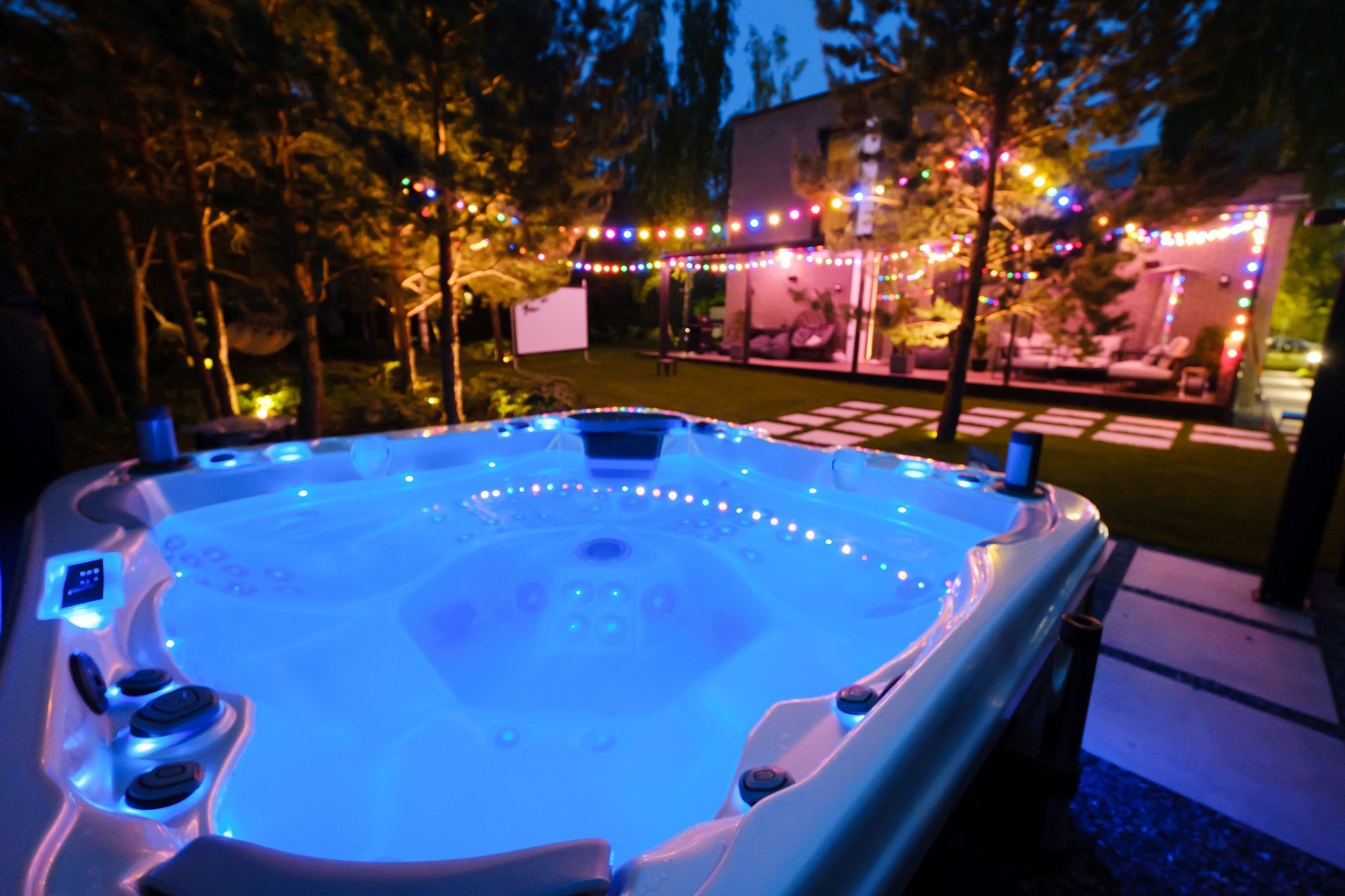 Un bain à remous illuminé de bleu sur une terrasse de jardin au crépuscule, avec des guirlandes lumineuses et une maison en arrière-plan