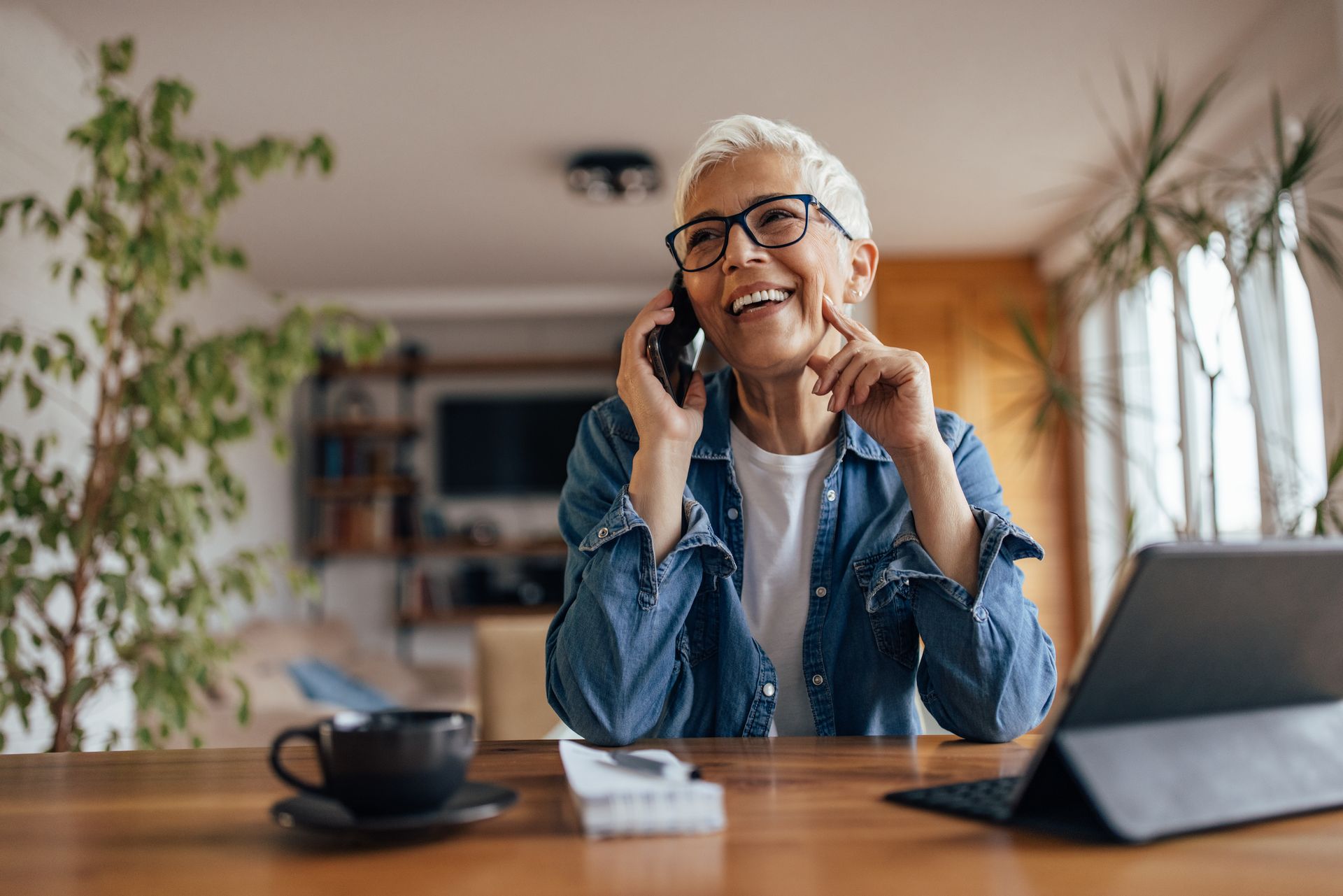 Personne souriante au téléphone, assise à une table de cuisine avec un ordinateur portable et une tasse de café