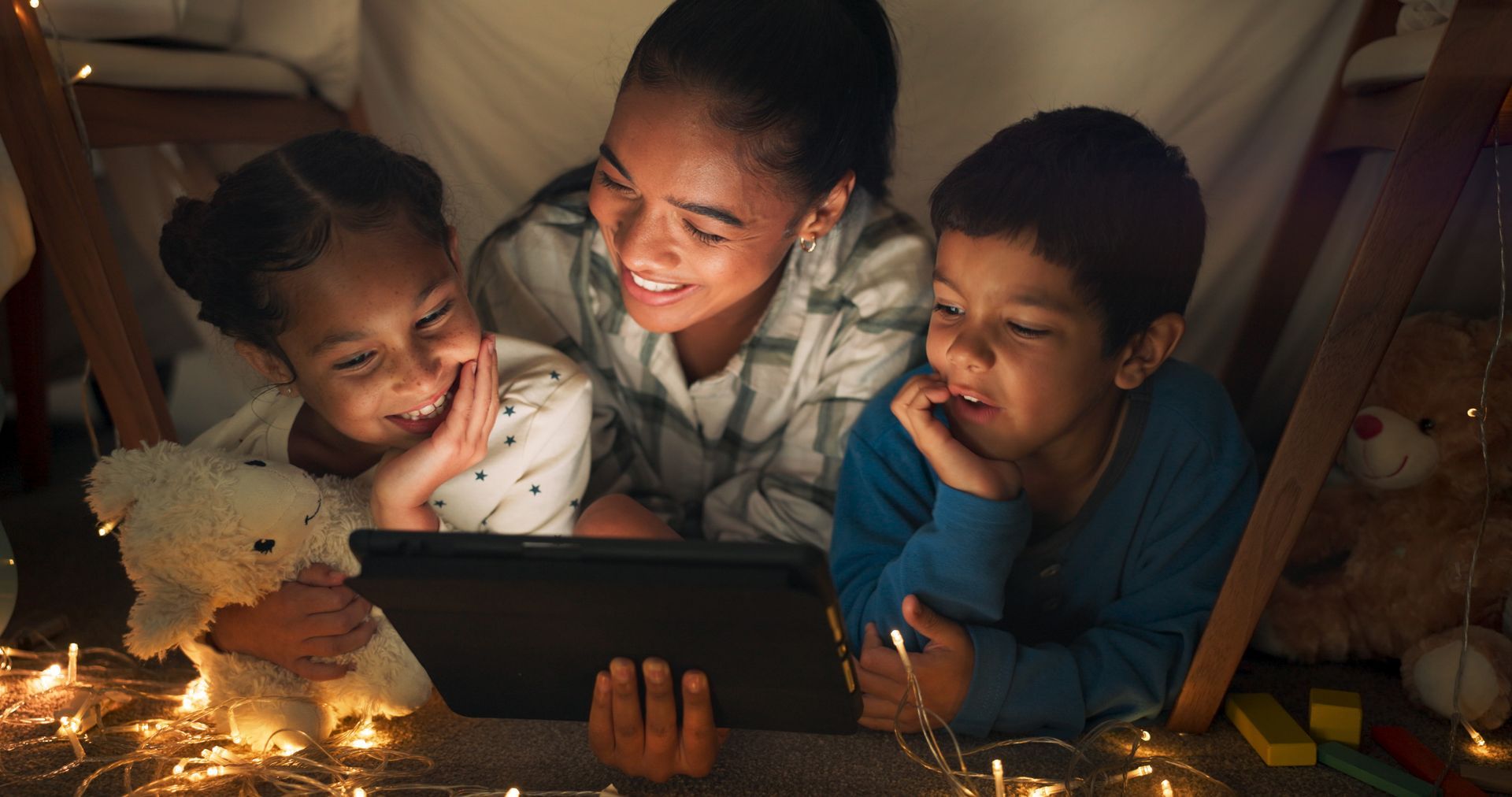 Trois enfants sont blottis autour d’une tablette dans une cabane douillette faite de couvertures, souriant à la lumière chaleureuse de guirlandes lumineuses.