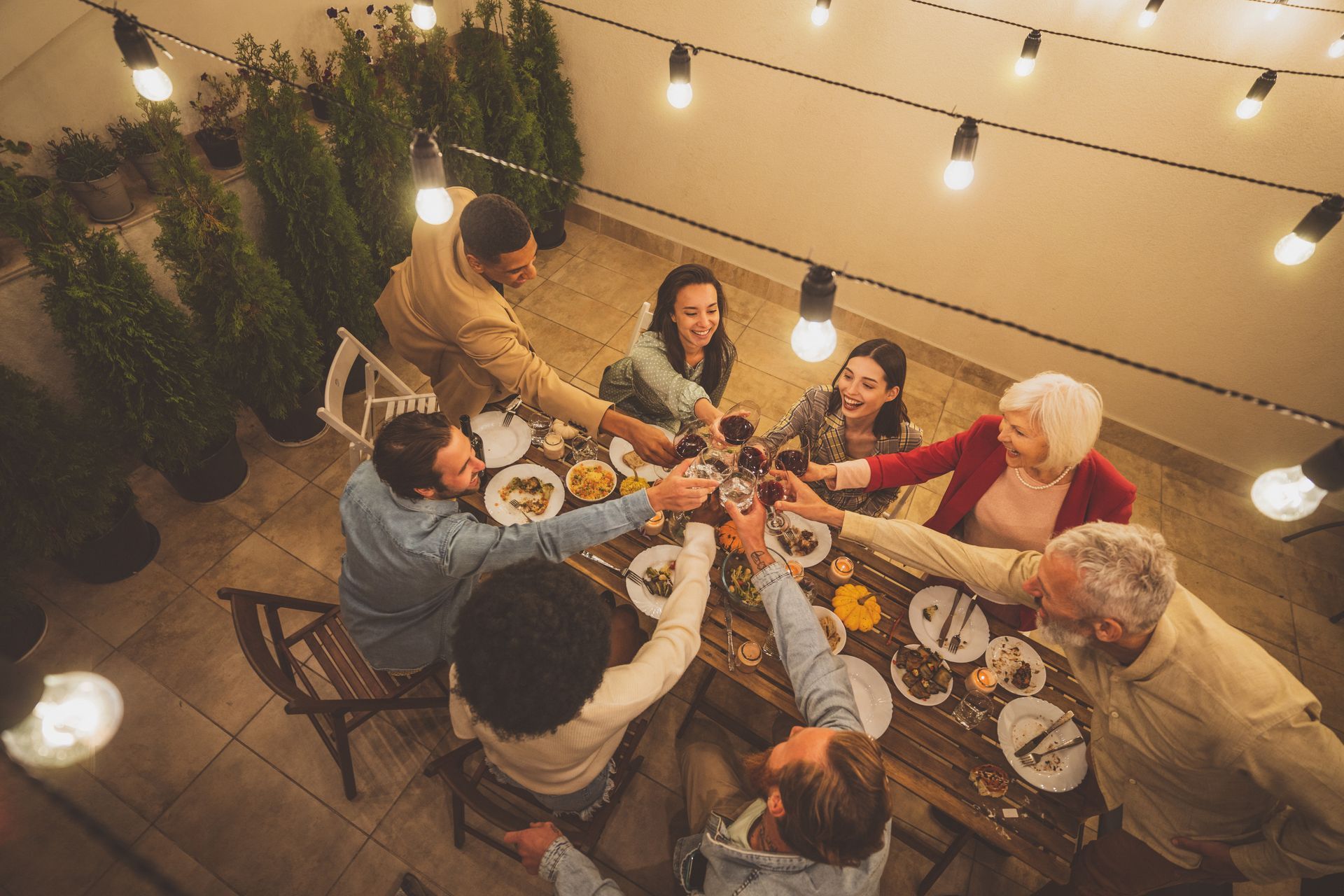 Des amis trinquent autour d’une table de fête, sous des guirlandes lumineuses chaleureuses, à la nuit tombée