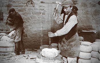 Two men in workshop carving curling stones with hammers and chisels.