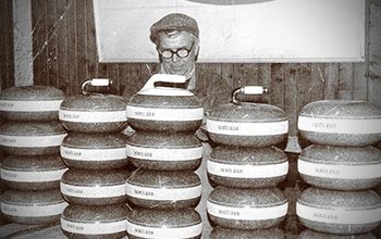 Man in cap and glasses surrounded by stacked curling stones in a wooden setting.