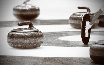 Curling stones on ice, some clustered near a target circle, with a curling brush in view.