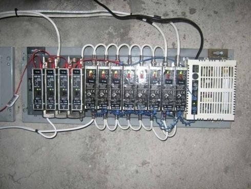 Un quadro elettrico di controllo grigio con diversi relè verticali e un alimentatore ventilato bianco montato a parete.