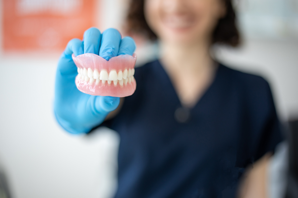 Dentist in blue gloves holding a set of dentures, smiling in an office setting.
