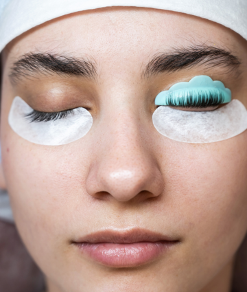Woman with closed eyes, undergoing a lash lift procedure, with a tool and eye pads on her eyelids.